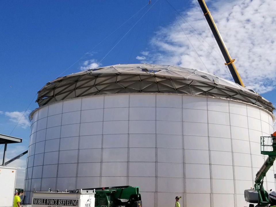 Aluminum dome roof being craned into place atop a white bolted steel tank