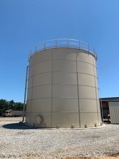 Completed tan bolted steel tank with a perimeter roof handrail on a gravel pad