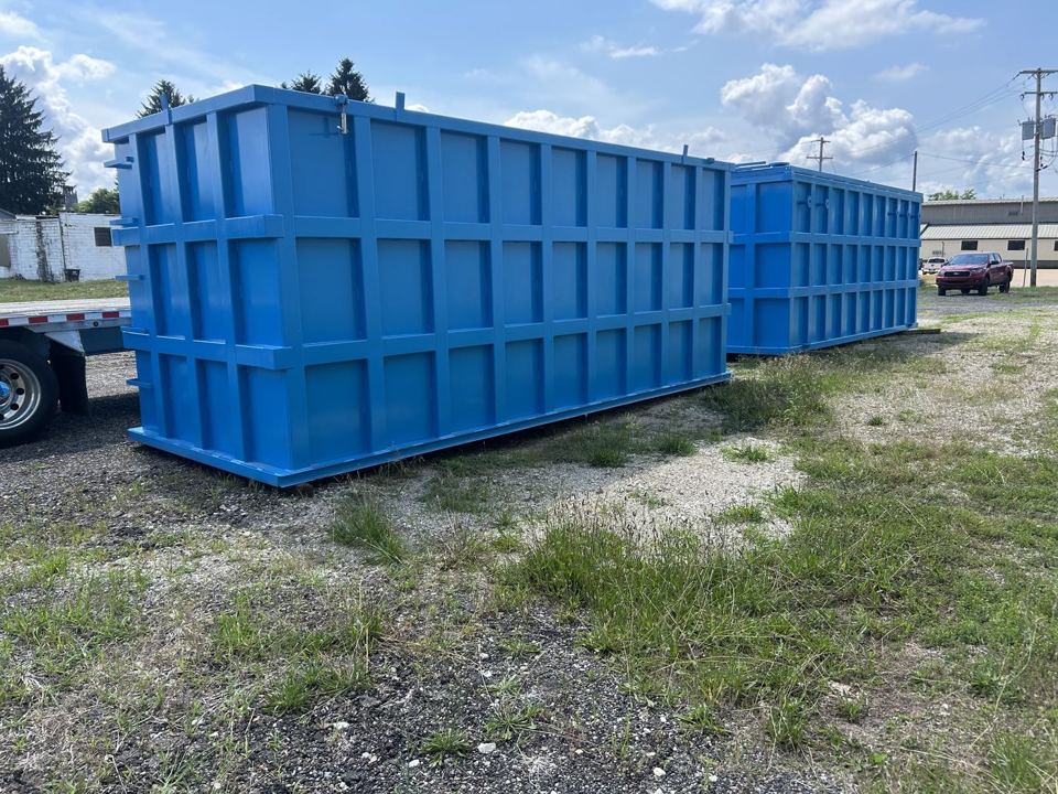 Pair of bright blue open-top shop-welded steel tanks staged side by side in a grassy yard, externally reinforced panel walls under a clear sky