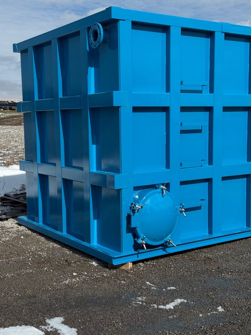 Corner of a blue shop-welded steel tank with a bolted, gasketed manway and wing-nut clamps, staged on a winter lot