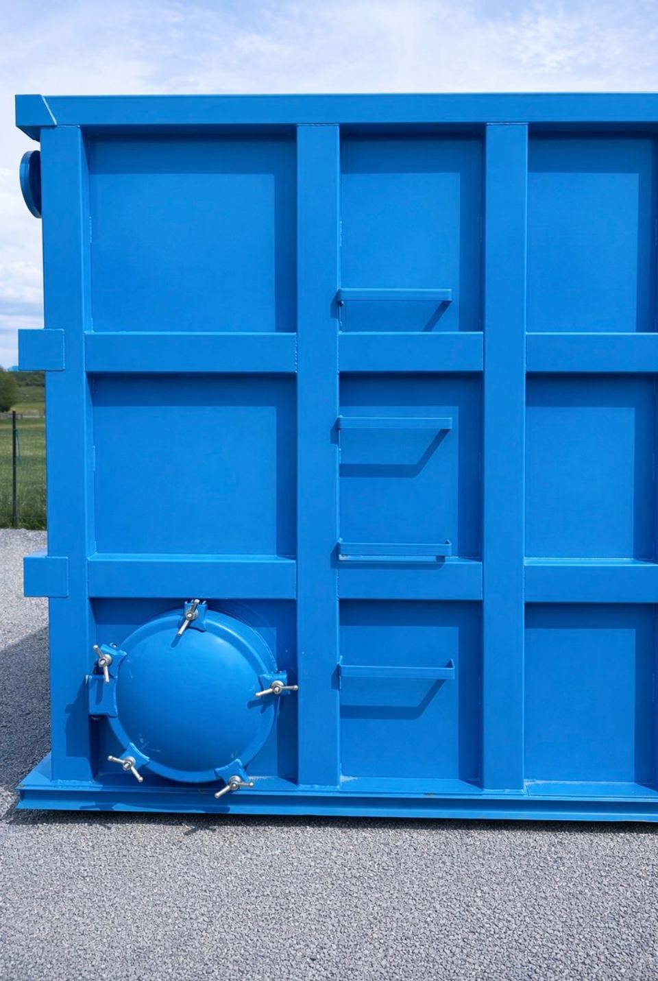 End wall of a blue shop-welded steel tank with a bolted circular manway and welded step brackets, outdoors on gravel