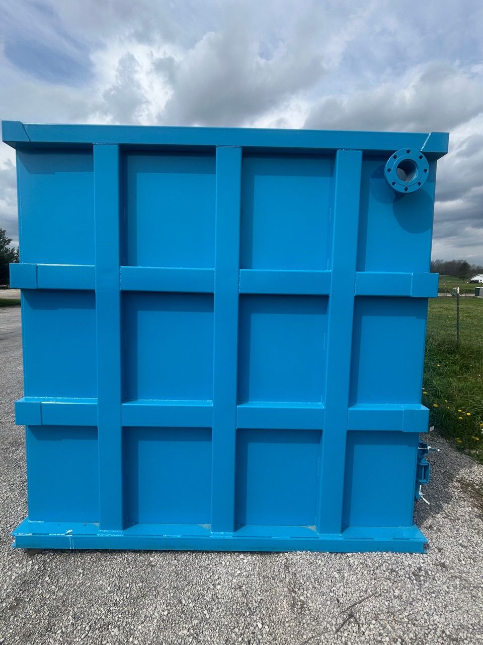 End wall of a finished blue shop-welded steel tank with its reinforced panel grid and a corner flanged nozzle, outdoors under a cloudy sky