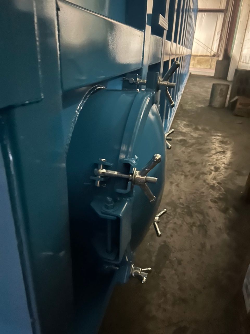 Close-up of a side-mounted bolted manway with swing-bolt wing-nut clamps on a blue shop-welded steel tank in the shop