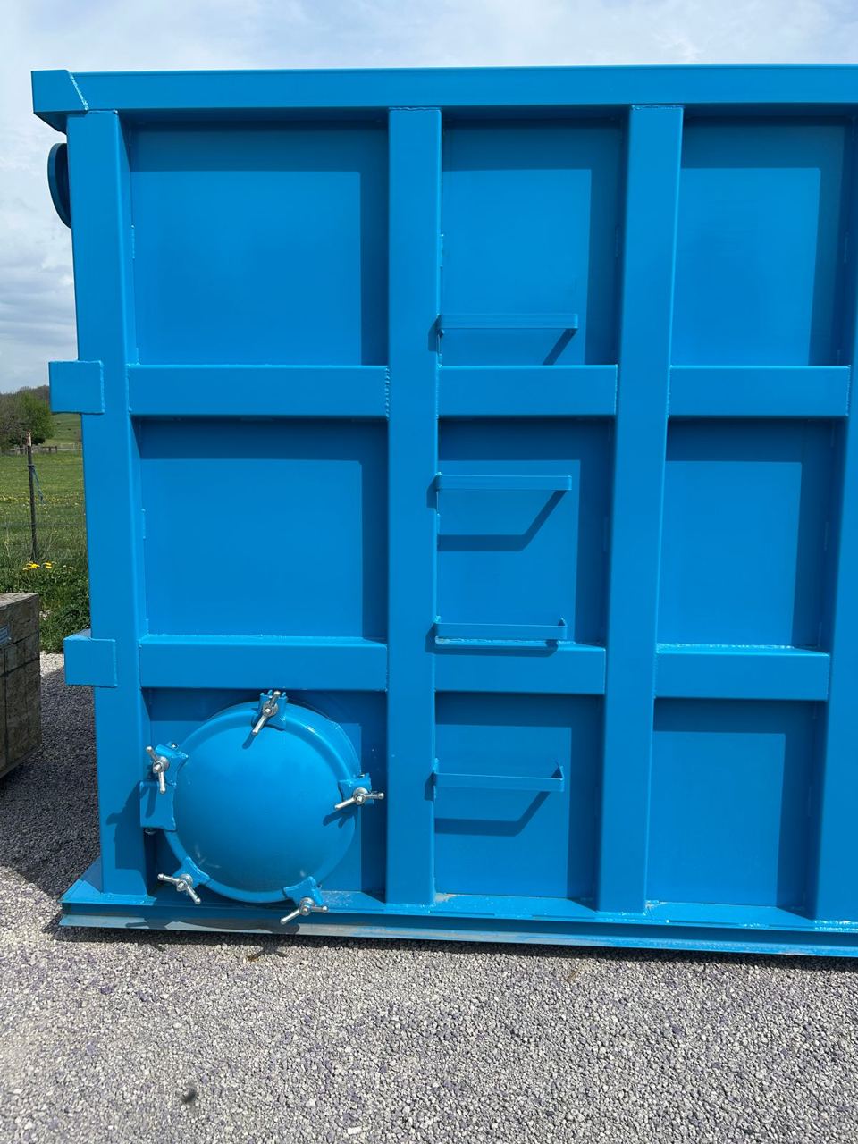 End wall of a blue shop-welded steel tank with a bolted manway and welded step brackets for access, staged outdoors