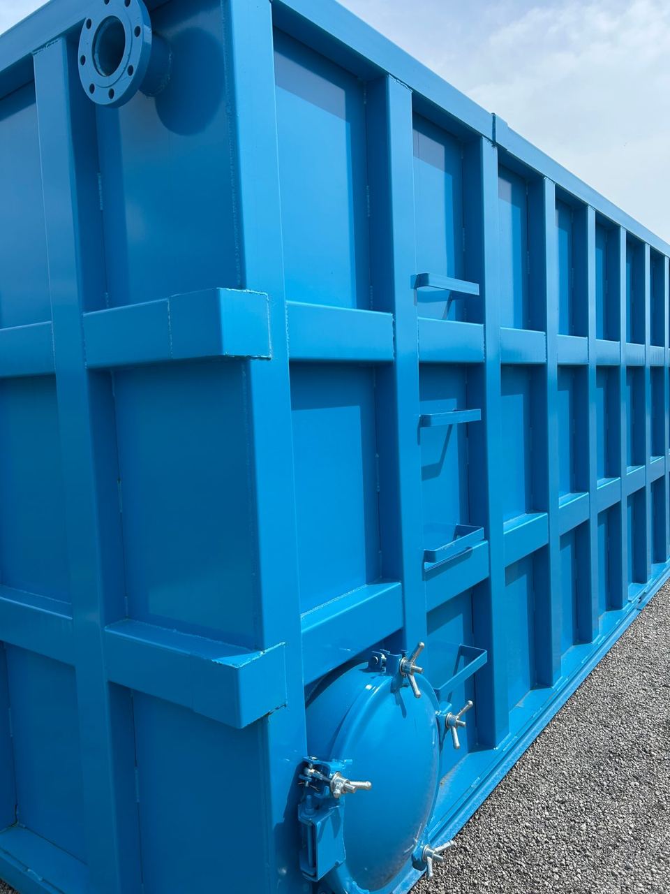 Long side of a blue shop-welded steel tank outdoors showing the reinforced panel grid, top nozzle, and a side-mounted bolted manway