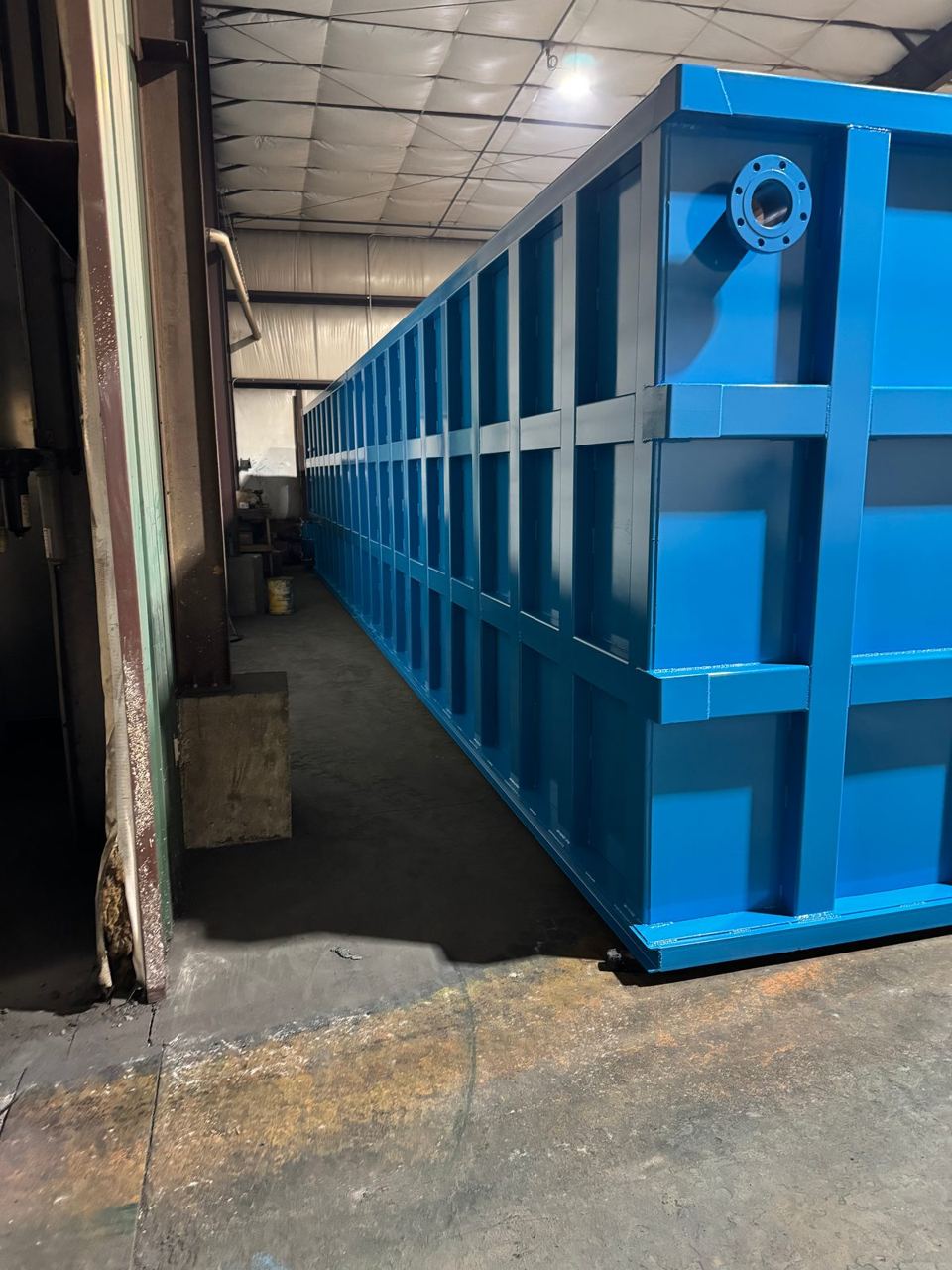 Blue shop-welded steel tank running down a narrow shop aisle, top-corner flanged nozzle in the foreground
