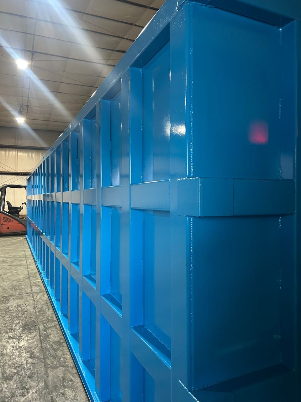 Freshly coated blue shop-welded steel tank running the length of the fabrication shop, glossy panels reflecting the overhead lights