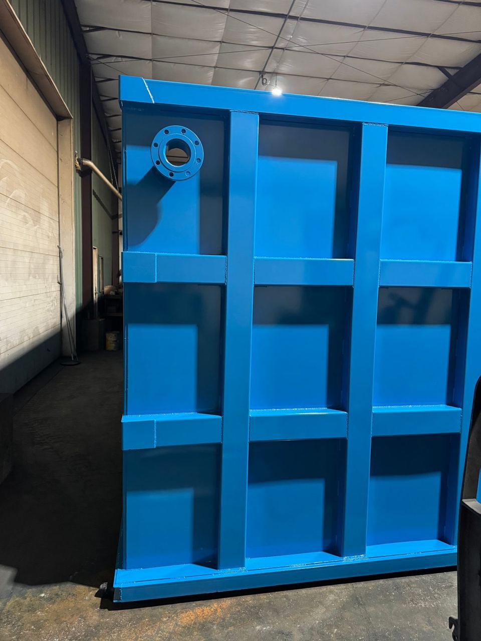 Tall blue shop-welded steel tank end section with a flanged nozzle and three-by-three reinforced panel grid inside the fabrication shop