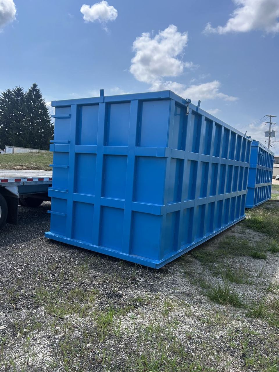 Three-quarter view of a single bright blue modular steel tank staged outdoors on gravel, reinforced panel grid and a flatbed trailer alongside