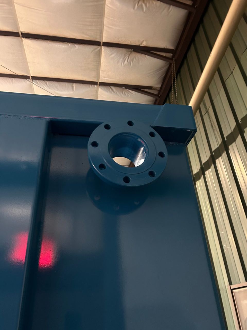 Detail of a bolt-pattern flanged nozzle welded to the top corner of a dark blue modular steel tank in the shop