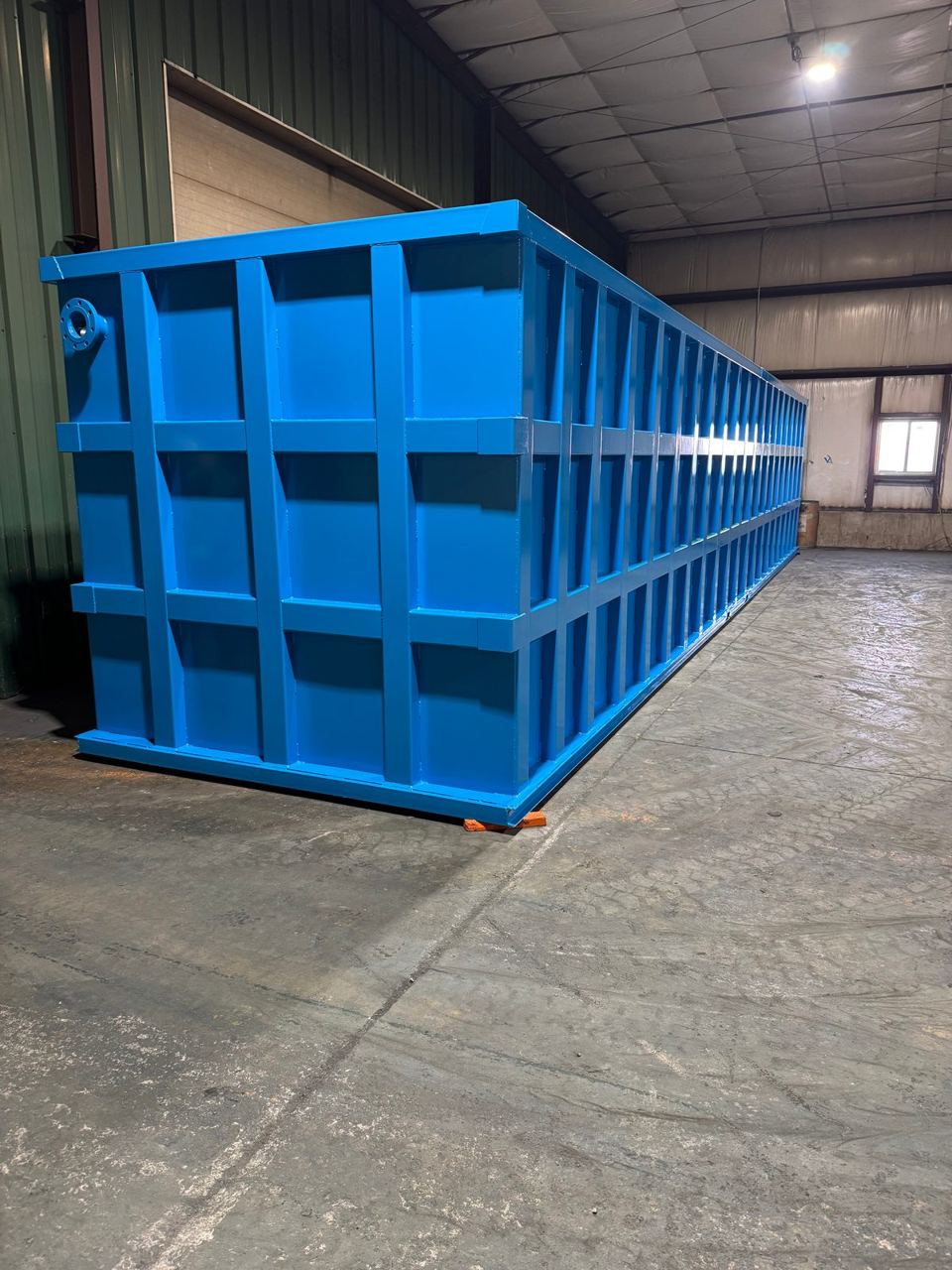 Three-quarter view of a long blue modular steel tank in the shop with a welded lifting lug at the corner