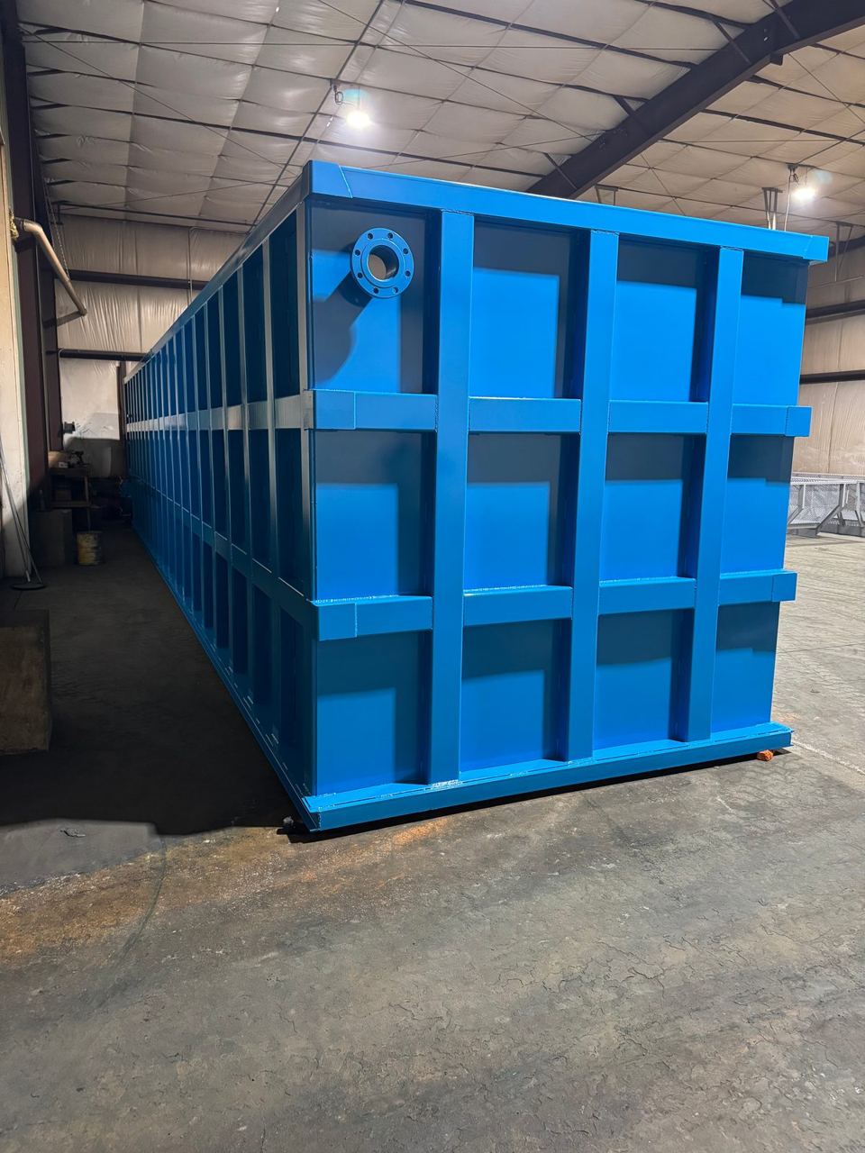 Long blue modular steel tank in the shop with a top-corner flanged nozzle and the reinforced panel grid running its full length