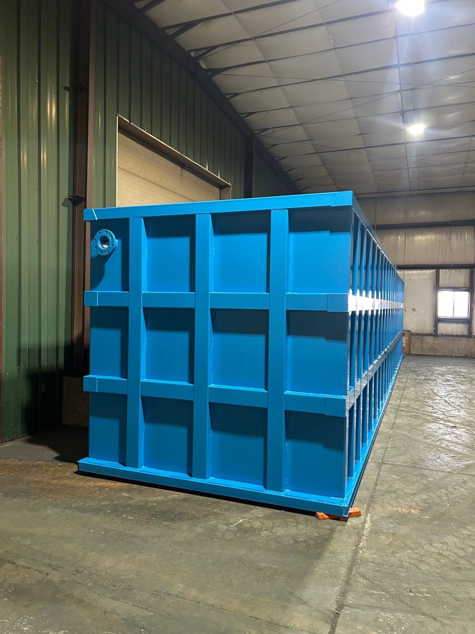 Three-quarter view of a long blue modular steel tank inside the shop, lifting lug on the near corner
