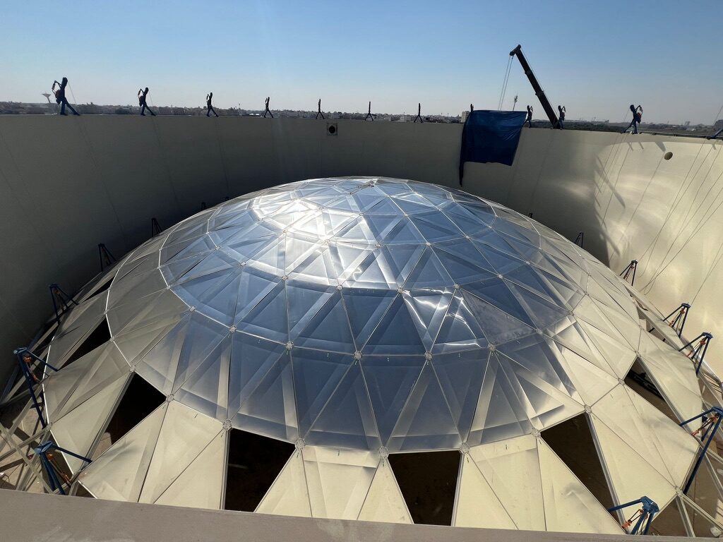 Aluminum geodesic dome cover under installation on top of a large white storage tank