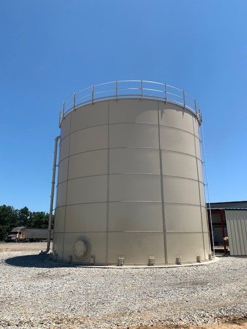 Completed tan bolted steel tank with a perimeter roof handrail on a gravel pad