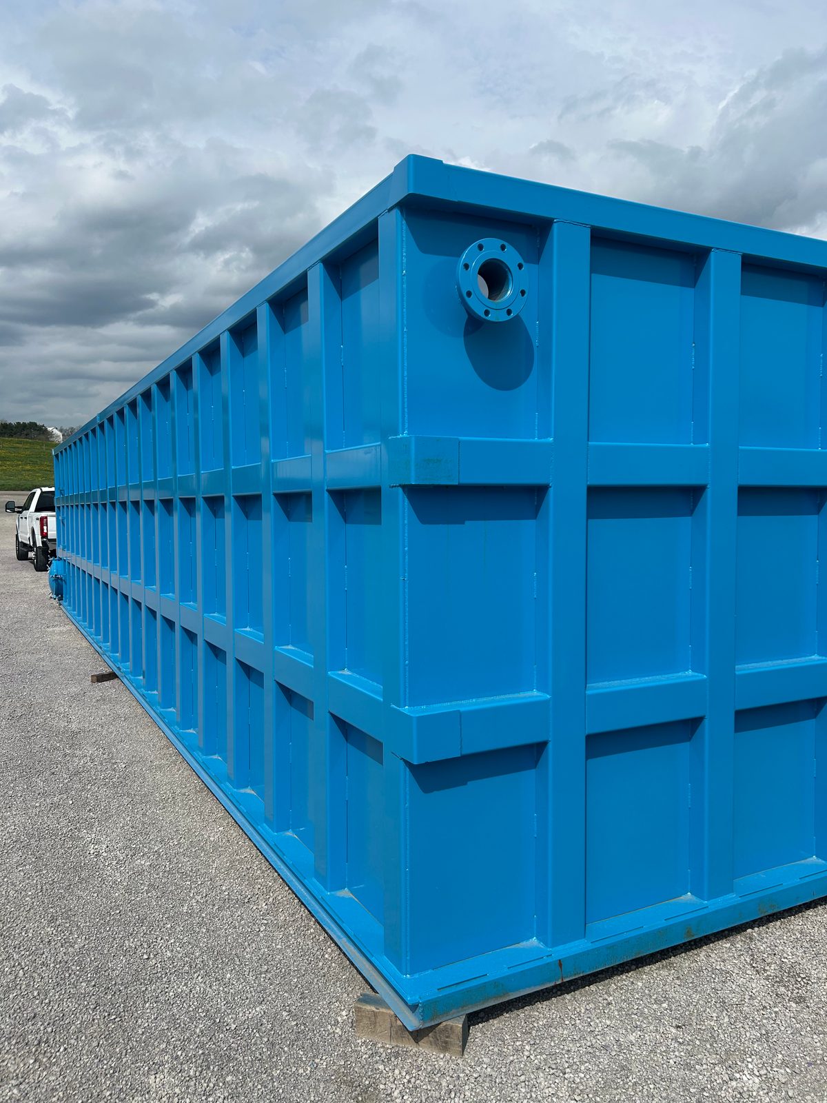 Large blue modular steel tank staged outdoors with a pickup truck alongside for scale, top nozzle and bolted panel seams