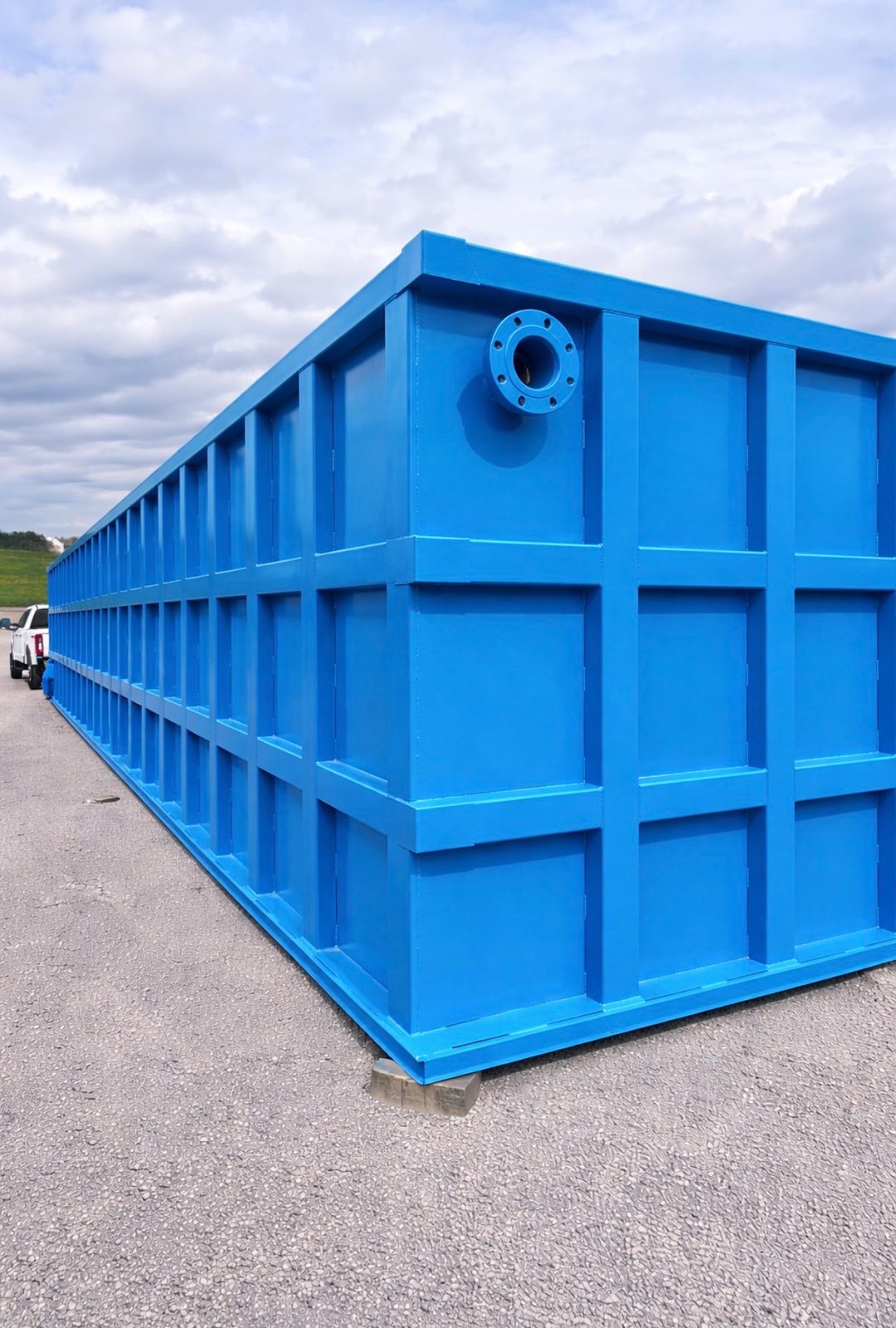 Long blue modular steel tank staged outdoors in the yard, top-corner flanged nozzle and reinforced panel grid receding to the horizon