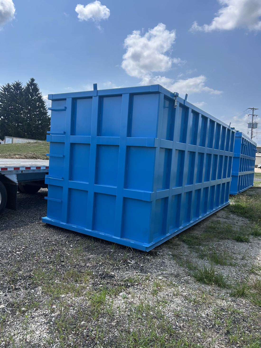 Three-quarter view of a single bright blue modular steel tank staged outdoors on gravel, reinforced panel grid and a flatbed trailer alongside