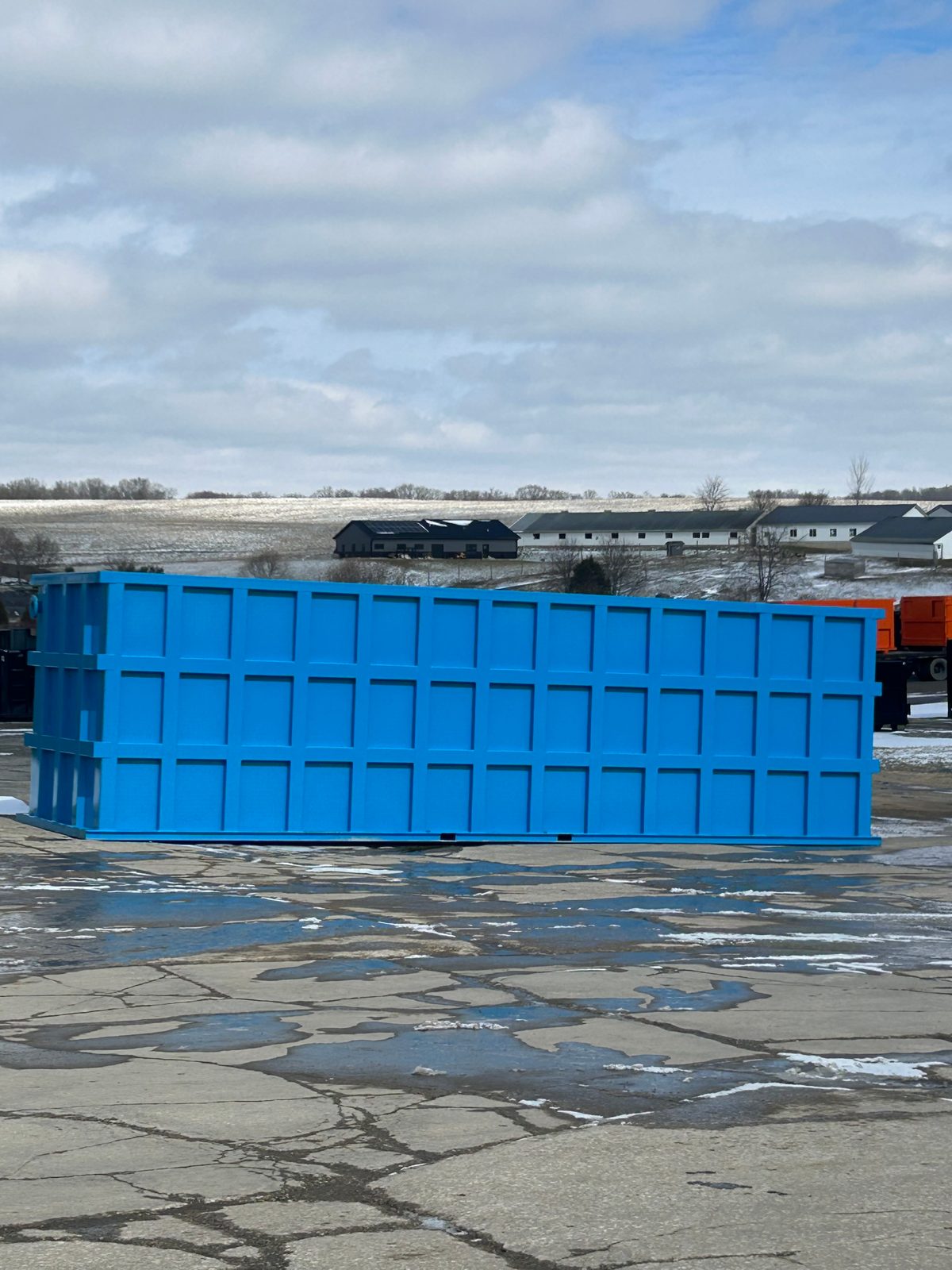 Blue modular steel tank staged on a snow-dusted yard with farm buildings on the horizon